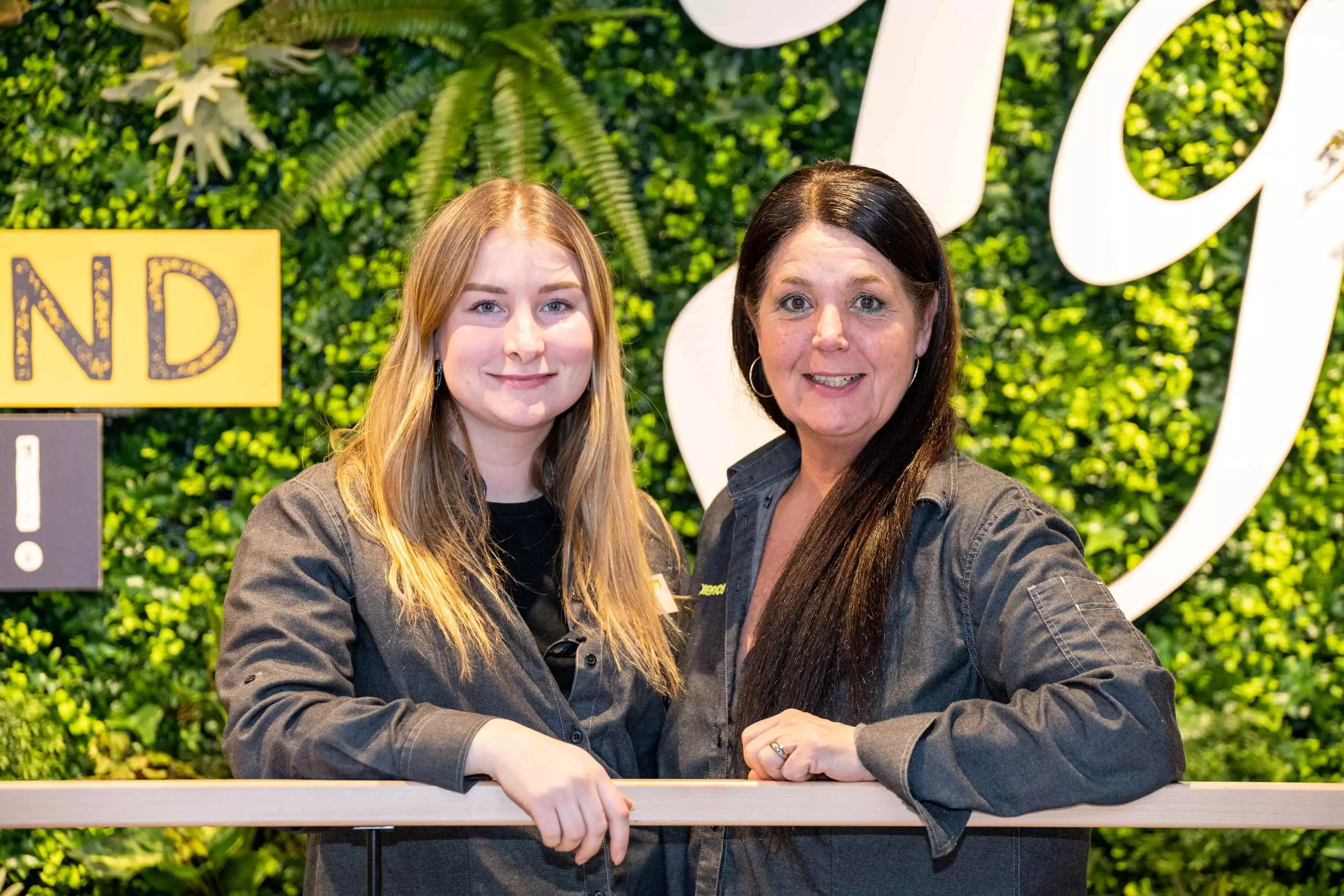 "Jonge vrouw met zwart, krullend haar in een staart glimlacht terwijl ze op een kassa leunt. De toonbank heeft een klein plantje in een pot en een pinapparaat. De winkel heeft houten wanden en kantoorverlichting."